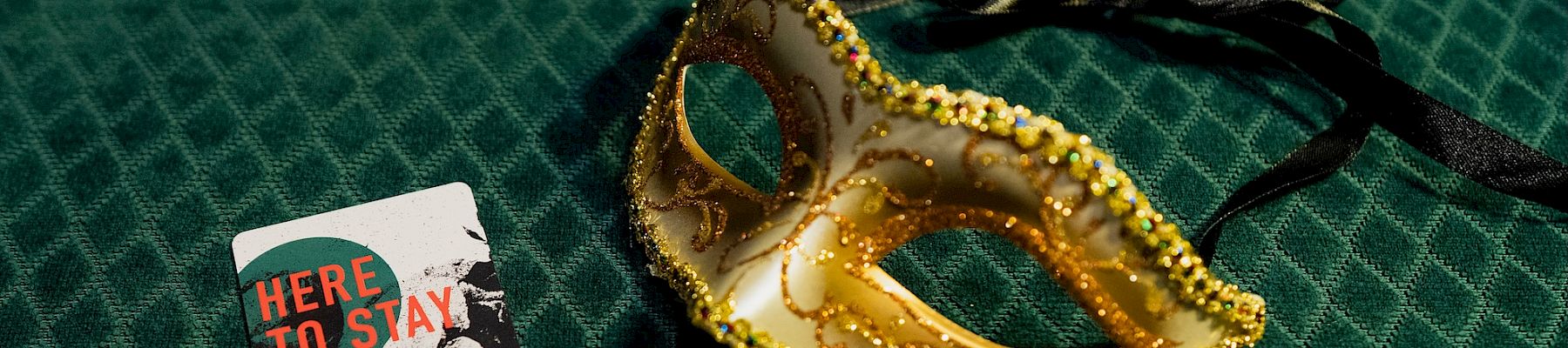 A gold masquerade mask with black ribbons beside a small card that says &ldquo;HERE TO SLAY&rdquo; on a green textured surface.