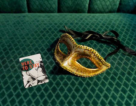 A gold masquerade mask with black ribbons beside a small card that says &ldquo;HERE TO SLAY&rdquo; on a green textured surface.