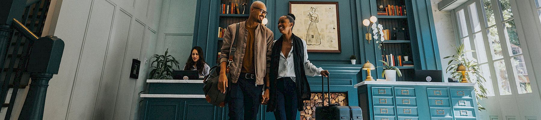 Two travelers stand with a suitcase in a stylish teal hotel lobby, smiling as they check in or chat near the reception.