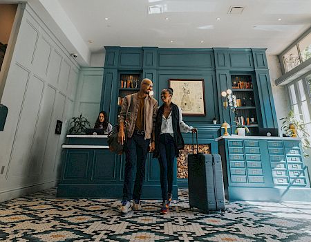 Two travelers stand with a suitcase in a stylish teal hotel lobby, smiling as they check in or chat near the reception.