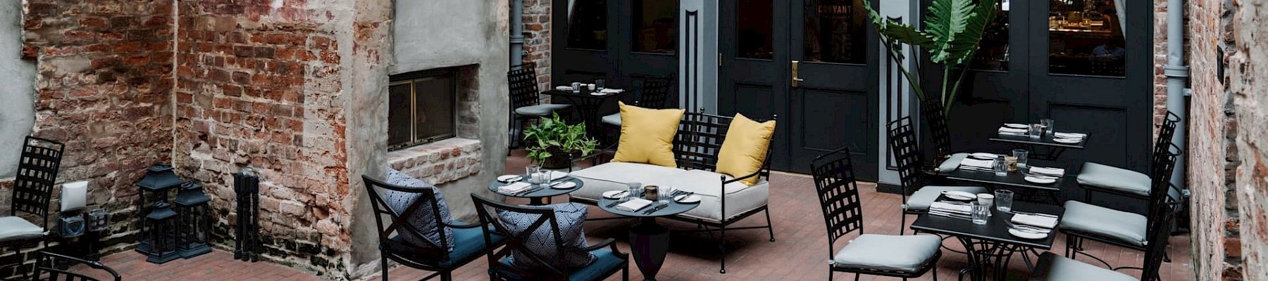 A cozy urban courtyard cafe with exposed brick walls, black-framed doors, and neatly arranged tables and chairs outdoors.
