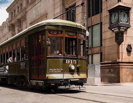 A vintage streetcar named &ldquo;915&rdquo; travels along a rail line beside a historic stone building and a classic clock lantern on a sunny city street.