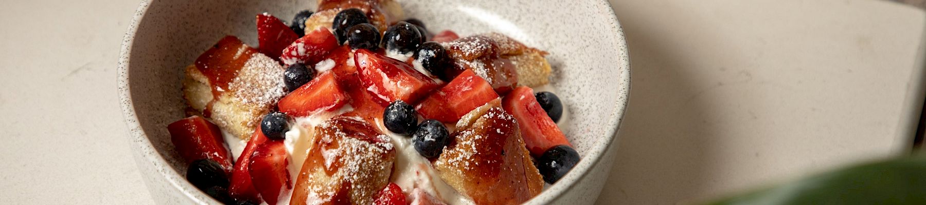 A bowl of dessert with whipped cream, topped with chunks of bread or cake, strawberries, blueberries, and a dusting of powdered sugar.