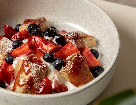 A bowl of dessert with whipped cream, topped with chunks of bread or cake, strawberries, blueberries, and a dusting of powdered sugar.