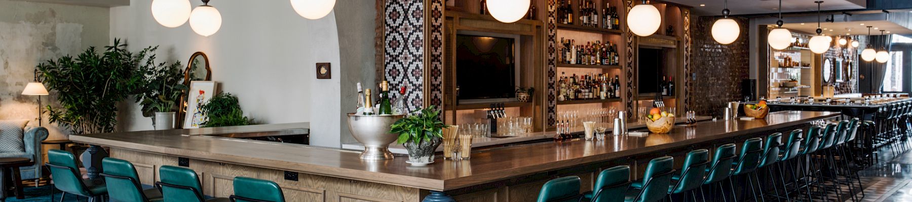 A stylish, long wooden bar with teal stools, checkered floor, round pendant lights, plants, and shelves behind the bar.