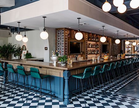 A stylish, long wooden bar with teal stools, checkered floor, round pendant lights, plants, and shelves behind the bar.