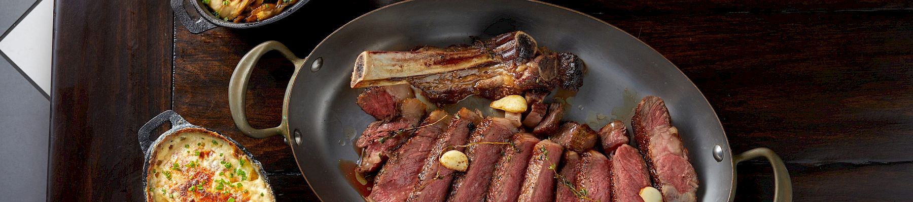 A sizzling steak platter with sliced beef, grilled bone-in rib, garlic butter, plus two side dishes and a sauce boat on a dark wooden table, delicious.
