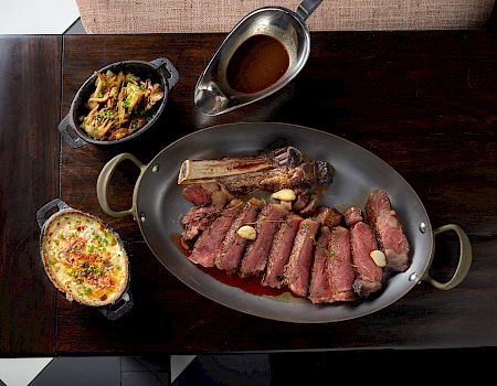 A sizzling steak platter with sliced beef, grilled bone-in rib, garlic butter, plus two side dishes and a sauce boat on a dark wooden table, delicious.