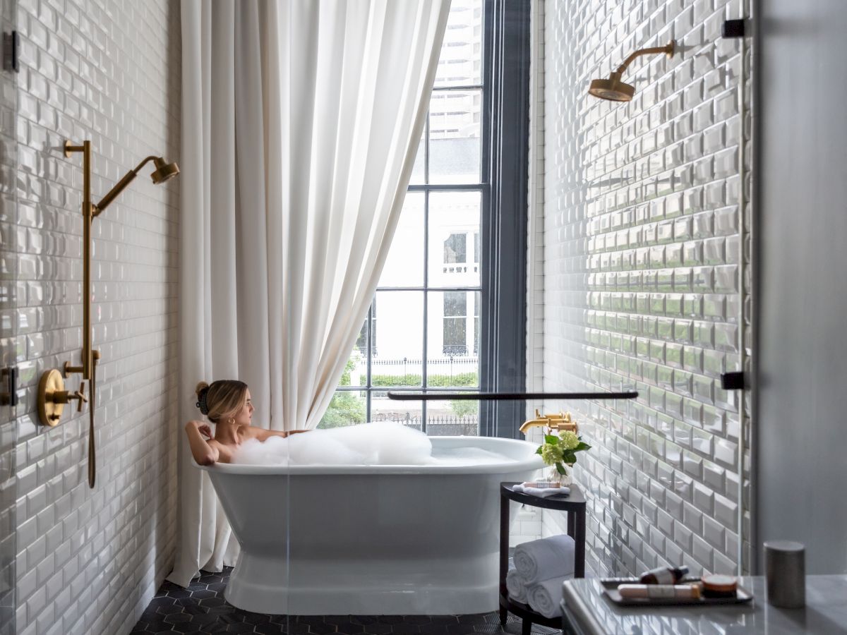 A person relaxes in a white freestanding bathtub by a large window, soft curtains, brass wall lamps, and a small side table with flowers. Ends with a period.