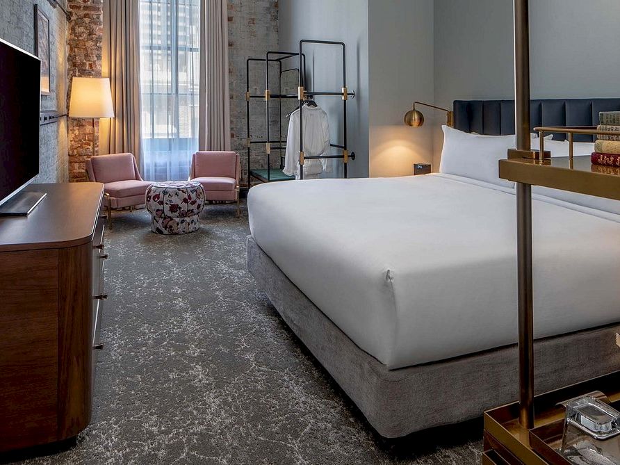 A spacious hotel room with a large bed, stylish seating area, flat-screen TV on a wooden dresser, and a window letting in soft light.