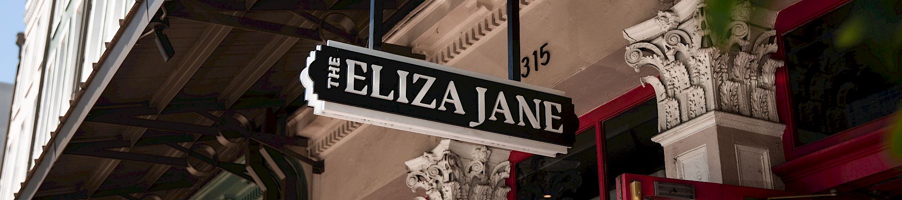 A storefront with ornate columns and a hanging sign reading &ldquo;FELIZA JANE&rdquo; above red doors, partly framed by greenery.