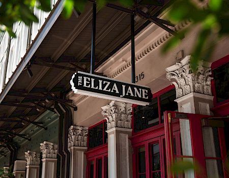 A storefront with ornate columns and a hanging sign reading &ldquo;FELIZA JANE&rdquo; above red doors, partly framed by greenery.