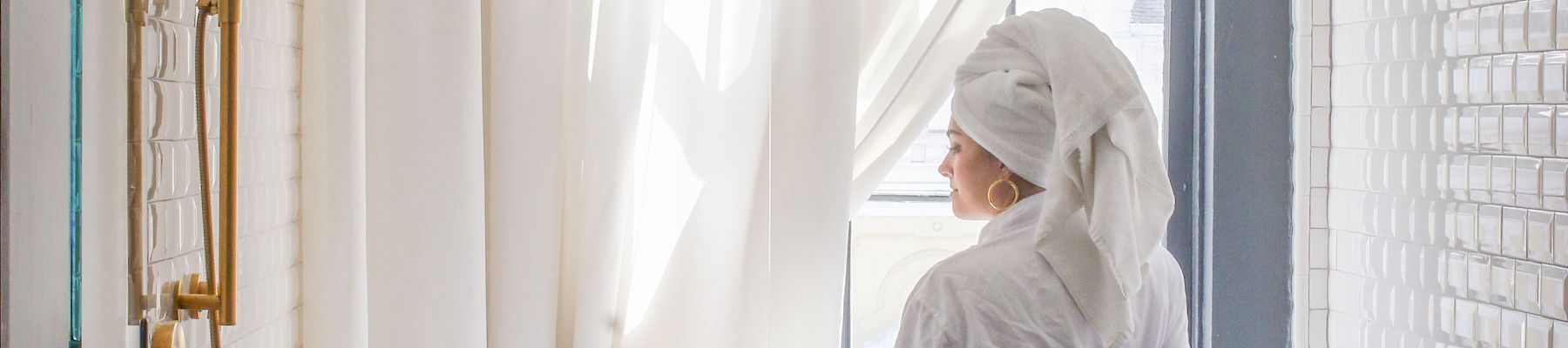 A person wearing a white robe sits at a freestanding bathtub in a bright bathroom with sheer curtains, gold fixtures, and a tiled floor.