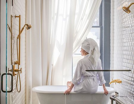 A person wearing a white robe sits at a freestanding bathtub in a bright bathroom with sheer curtains, gold fixtures, and a tiled floor.