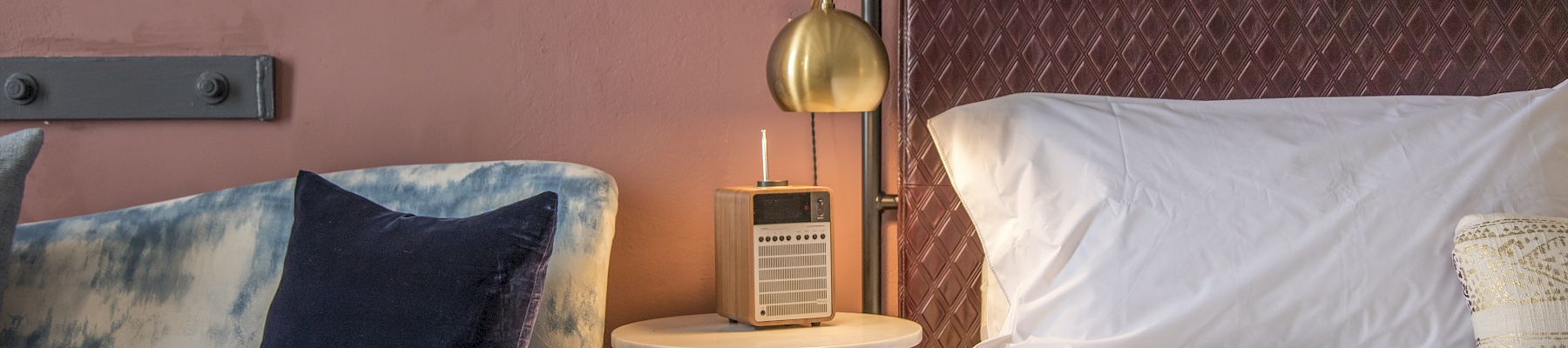 A cozy hotel room with a bedside table, lamp, and a plush bed flanked by a blue velvet couch, warm lighting, and soft decor. (Note: I kept it under 140 chars and ending with a period.)