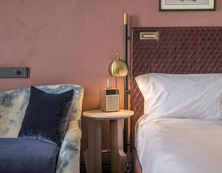 A cozy hotel room with a bedside table, lamp, and a plush bed flanked by a blue velvet couch, warm lighting, and soft decor. (Note: I kept it under 140 chars and ending with a period.)