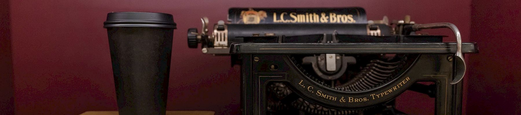 A vintage L.C. Smith & Bros typewriter sits on a red shelf beside a stack of books and a coffee cup.