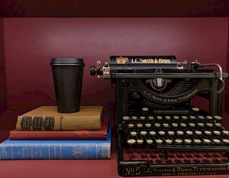 A vintage L.C. Smith & Bros typewriter sits on a red shelf beside a stack of books and a coffee cup.