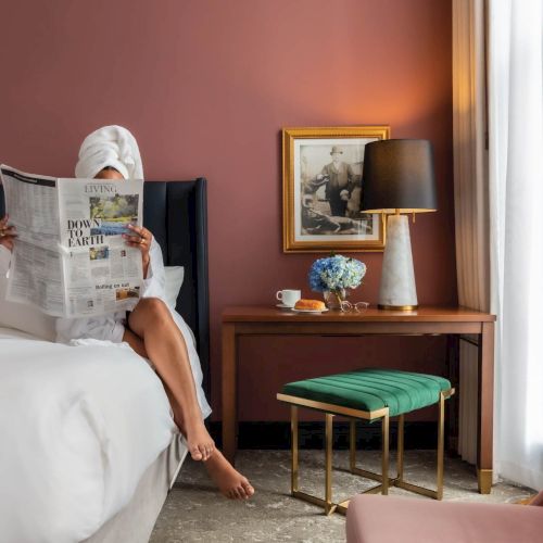 A cozy bedroom with a bed, a person sitting on it, warm lighting, a framed picture on the wall, and a small bedside table with a lamp.