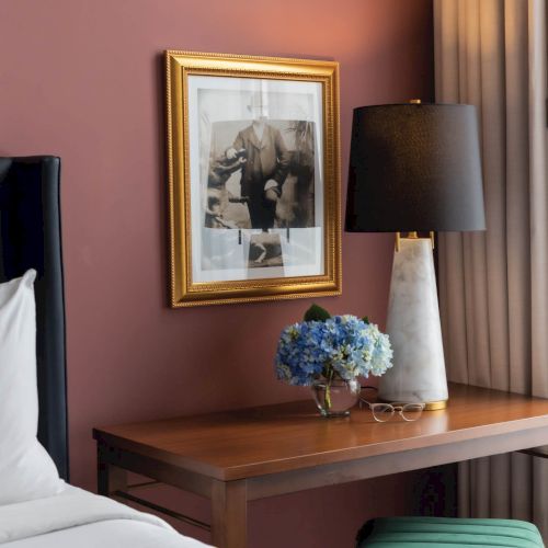 A cozy bedroom corner featuring a wooden dresser with a lamp, a framed artwork above it, and a small vase of blue flowers on the dresser.