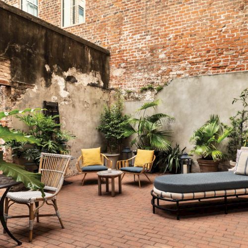 A small urban courtyard with brick flooring, potted plants, and outdoor lounge furniture including a chaise, chairs, and a table, creating a cozy retreat.