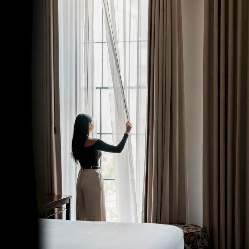 A woman stands by a tall window, pulling back sheer curtains as soft light fills the room, with a bed in the foreground and brown drapes framing the scene.