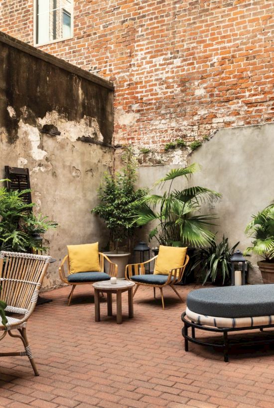 An urban rooftop garden with brick flooring, potted plants, lounge chairs, a cushioned daybed, and small tables along a brick wall.