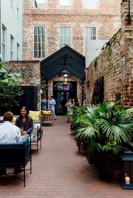 A cozy brick alley cafe with outdoor seating, leafy plants, and friends chatting under a sheltered archway at the end.
