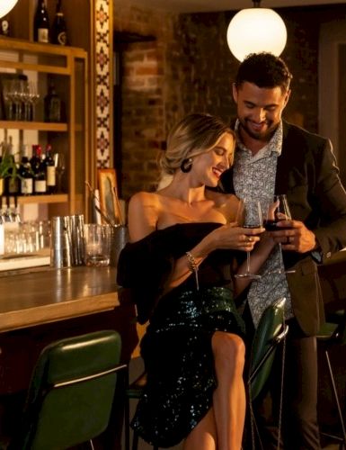Two stylish people toast and smile at a bar, while a bartender pours nearby, in a warm, dimly lit venue with vintage decor.