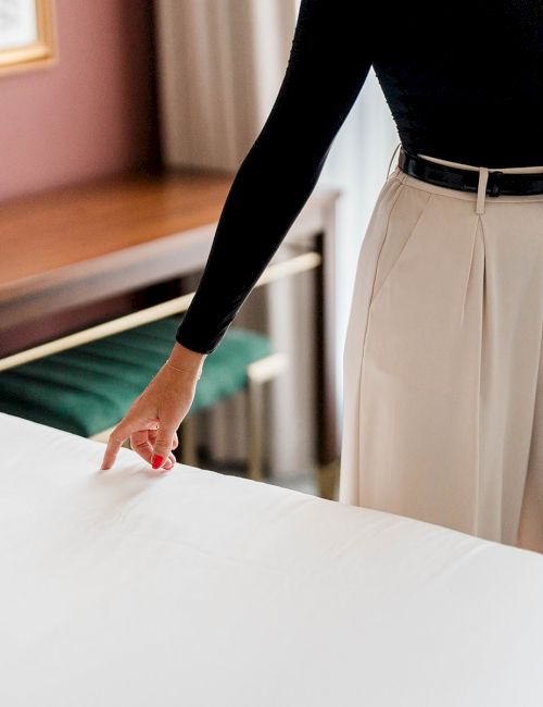 A person in a black top and light beige wide-leg pants, belt tied at the waist, standing by a bed and touching its white sheet.