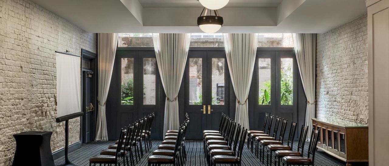A small, elegant conference room with brick walls, rows of black-and-white chairs facing a brick wall, and tall windows with white curtains ending the sentence.