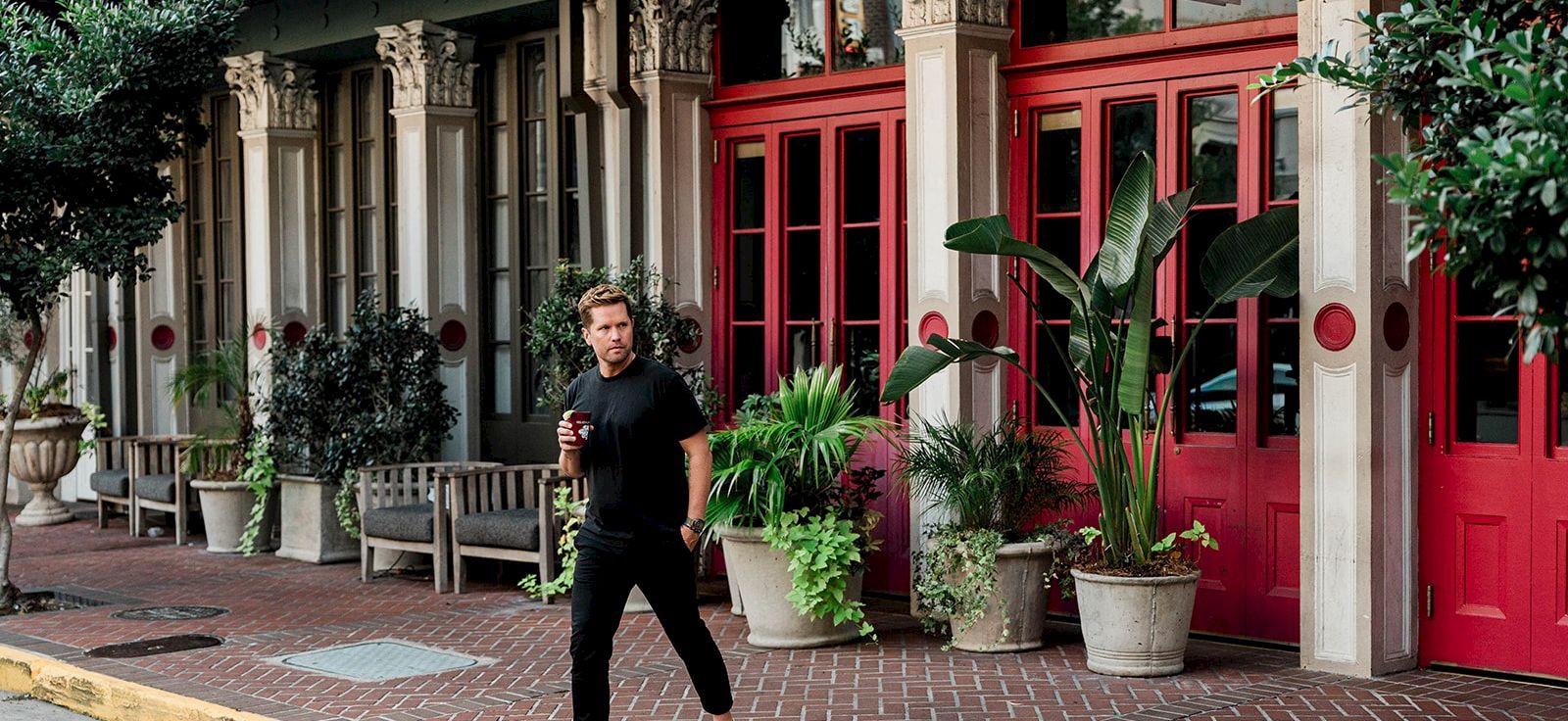 A man walking on a brick sidewalk past a row of bright red doors and ornate columns, with plants in pots by the storefronts, cheerful vibe.
