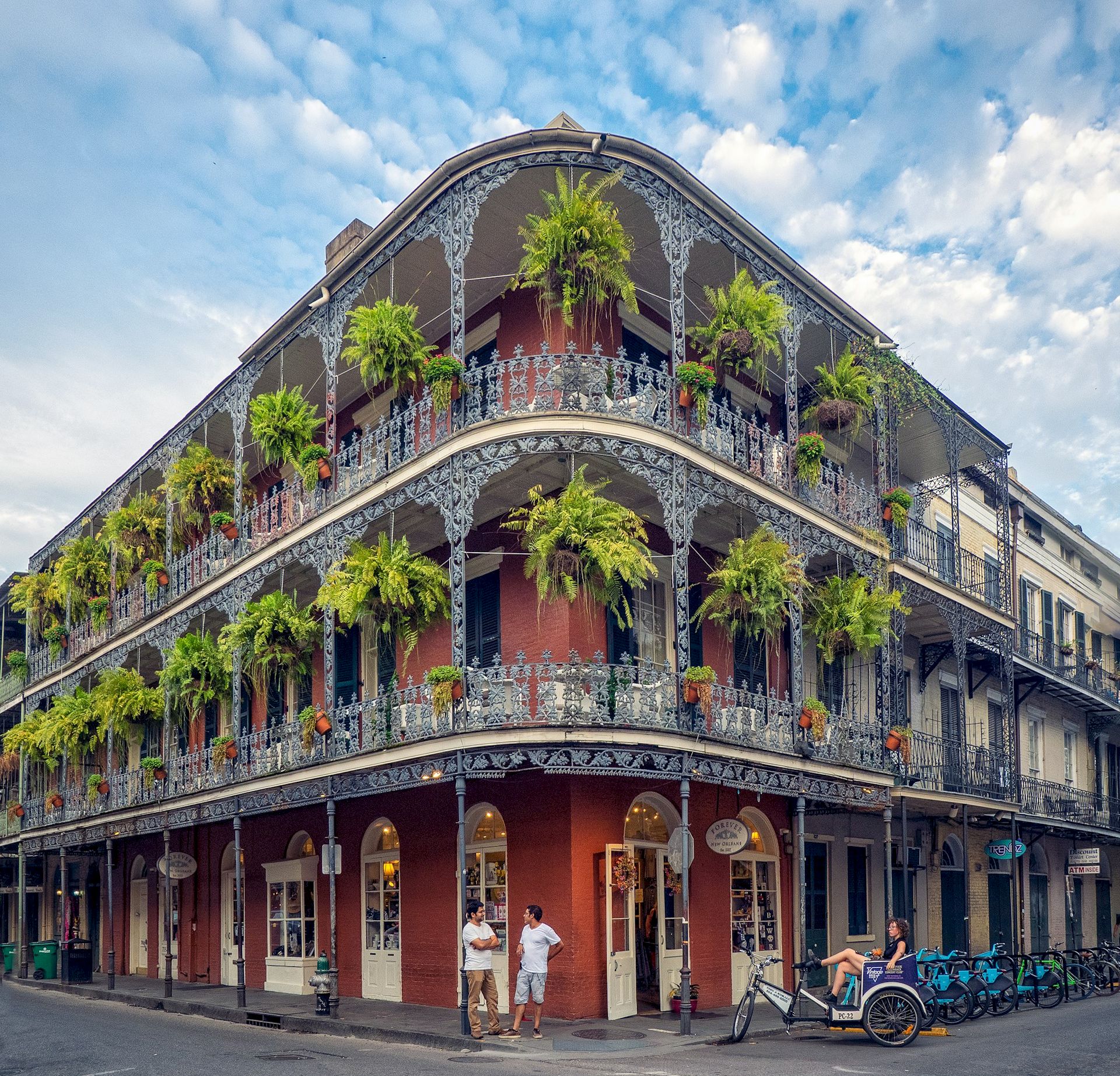 A vibrant corner building with ornate wrought-iron balconies, lush hanging plants, and colorful storefronts on ground level in a sunny street scene.