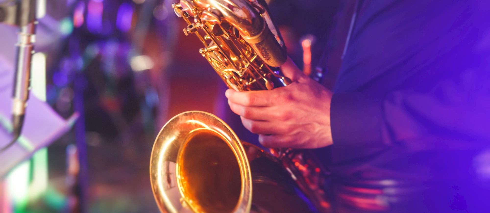 A close-up of a musician playing a saxophone on stage, fingers on keys, with colorful stage lighting in the background.