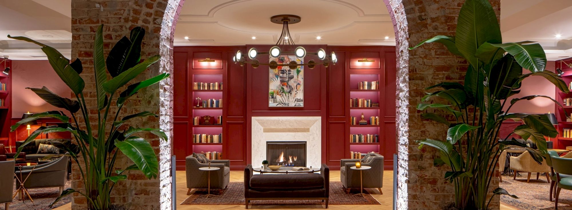 A cozy lounge with a brick archway, red wood shelves, a central fireplace, and lush green plants framing the seating area.