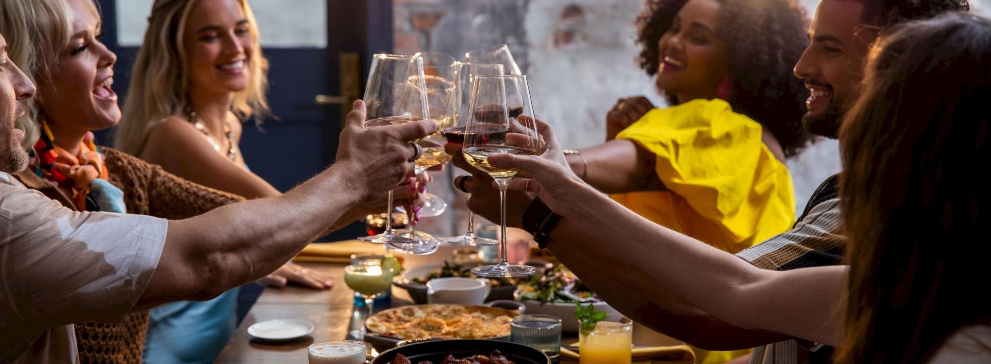 A group of friends toasting with wine glasses around a table, sharing food and good cheer at a restaurant or gathering.