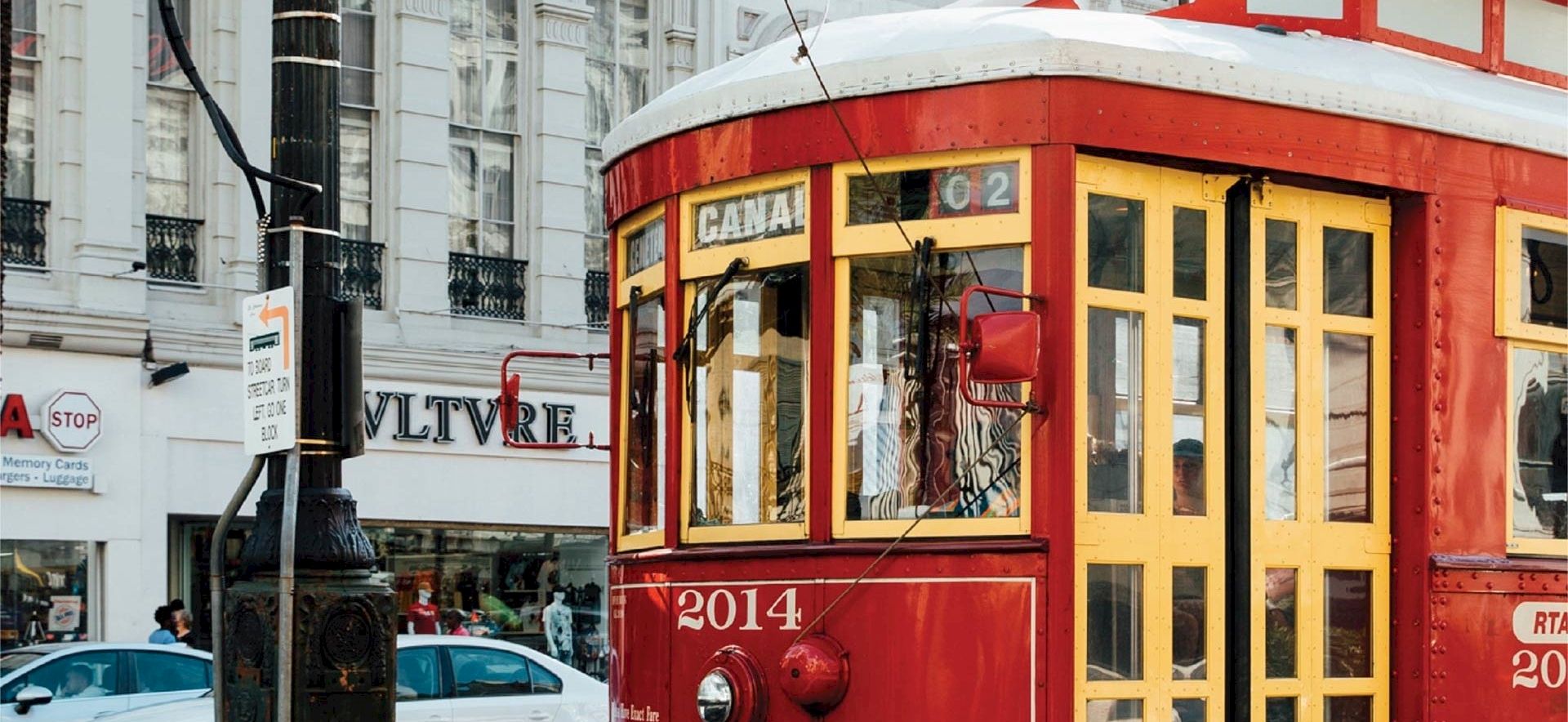 A red vintage tram on a city street with buildings and pedestrians, &ldquo;2014&rdquo; on its front, and a storefront sign in the background.