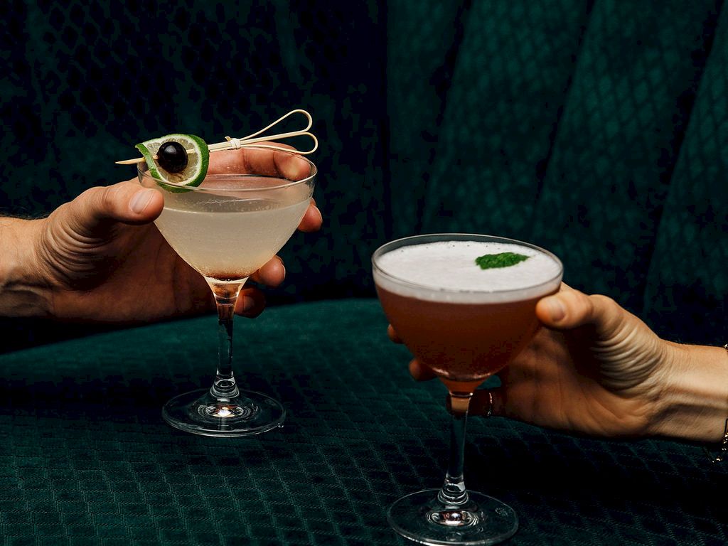 Two hands clink cocktail glasses, one with a lemon twist garnish and the other with a mint leaf, sharing a stylish, intimate toast.