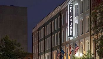 A row of brick townhouses at night with lit windows, a sign reading &ldquo;The Elizabeth Jane&rdquo; on the building, and American flags hanging outside.