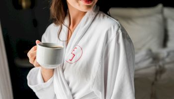 A person in a white robe holding a cup, standing indoors with a calm, relaxed pose and a partial smile.