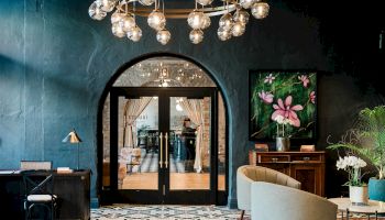 A stylish hotel lobby with a patterned floor, teal walls, arched glass doors, contemporary seating, and an overhead sculptural light fixture, inviting ambiance.