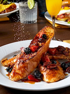 Delicious-looking French toast triangles with blueberries and syrup on a white plate, surrounded by drinks and other plates.