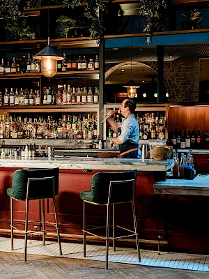 A bartender in a blue shirt pours a drink behind a red-bar with two green stools, shelves of bottles and warm hanging lights.