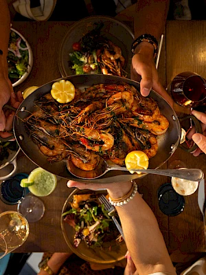 A table full of friends sharing a seafood feast, with a skillet of prawns and lemon wedges being passed around at a lively dinner.