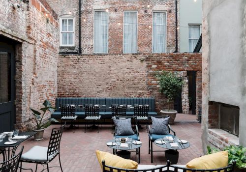 A cozy brick courtyard with a long black sofa, several chairs, small tables, potted plants, and a stone fireplace, all set in front of a brick building.