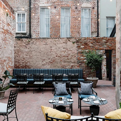 A cozy brick courtyard with a long black sofa, several chairs, small tables, potted plants, and a stone fireplace, all set in front of a brick building.