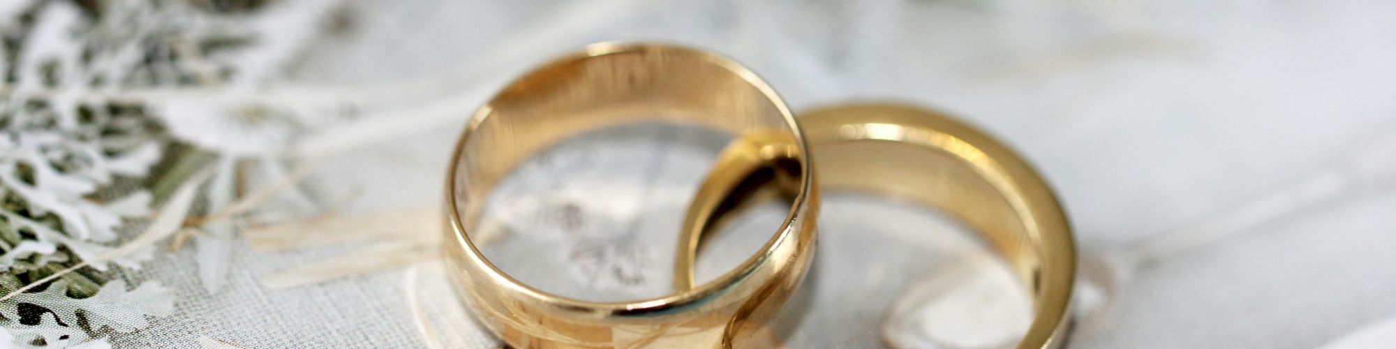 Two gold wedding bands rest on a light decor, with frosted branches and ribbon accents in the background, suggesting a wedding scene.