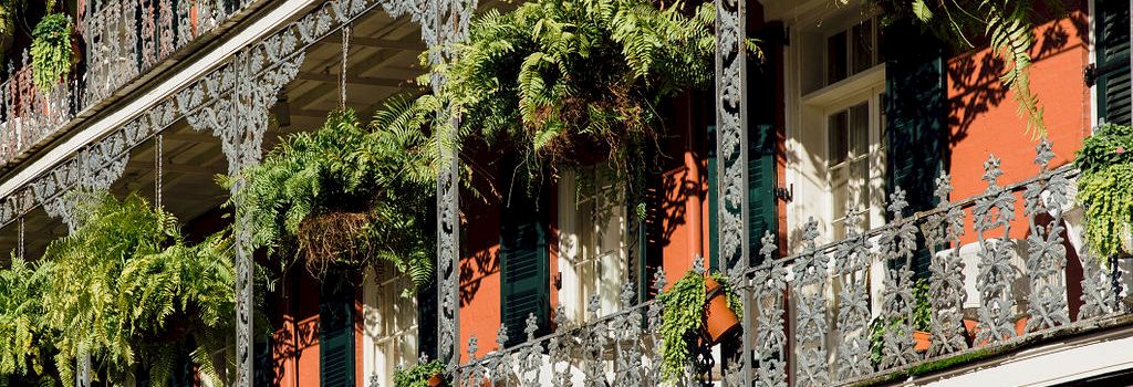 A cast-iron balcony with ornate railing drapes over a building, greenery hanging from planters, and red-orange walls beneath.