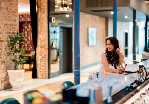 A stylish woman in a light dress sits at a bar counter in a modern, cozy cafe with brick walls and warm lighting, looking away.