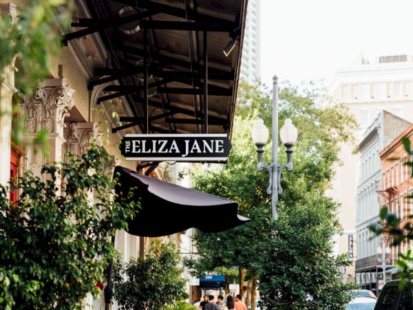 A street scene with greenery, storefronts, and a sign reading &ldquo;ELIZA JANE&rdquo; hanging above a sidewalk cafe or shop, bright day, urban vibe.